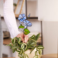 A person holding a stained glass representation of a morning glory plant stake, with blue flowers and green leaves, intended as table decor or a gift. Birth flower september.